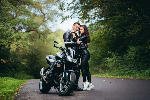 Handsome young man and beautiful young girl, couple sitting on a motorcycle, standing near a motorcycle in black leather clothes, hugging, in nature, on the street