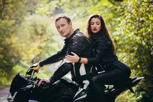 Handsome young man and beautiful young girl, couple sitting on a motorcycle in black leather clothes, close up, hugging, in nature, outdoors, looking at camera