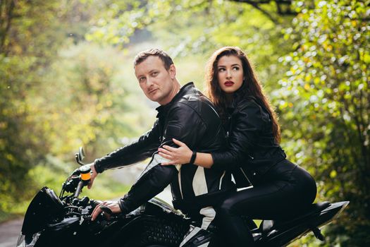 Handsome young man and beautiful young girl, couple sitting on a motorcycle in black leather clothes, close up, hugging, in nature, outdoors, looking at camera