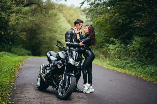 Handsome young man and beautiful young girl, couple sitting on a motorcycle, standing near a motorcycle in black leather clothes, hugging, in nature, on the street