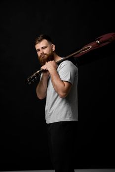 Young bearded guitarist in grey t-shirt holds a guitar on a shoulder and look in the camera in a dark room