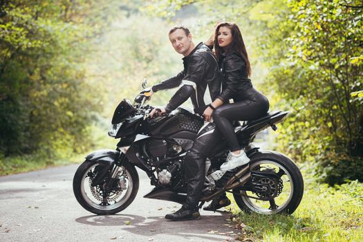 Handsome young man and beautiful young girl, couple sitting on a motorcycle, standing near a motorcycle in black leather clothes, hugging, in nature, on the street