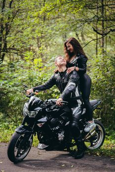 Handsome young man and beautiful young girl, couple sitting on a motorcycle in black leather clothes, close up, hugging, in nature, outdoors, looking at camera