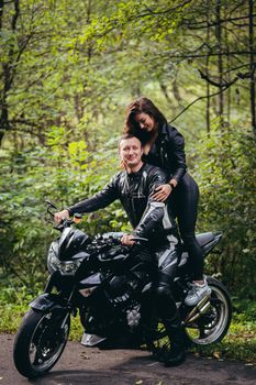 Handsome young man and beautiful young girl, couple sitting on a motorcycle in black leather clothes, close up, hugging, in nature, outdoors, looking at camera