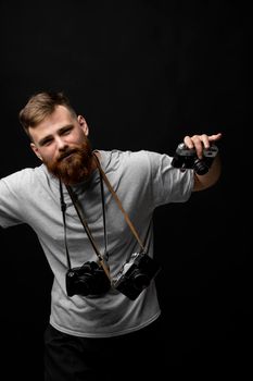 Handsome bearded photographer in a grey t-shirt with a bunch of different cameras in a hands and on a shoulder looking on a camera