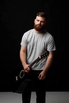 Young bearded guitarist in grey t-shirt holds a guitar in a hand in a dark room