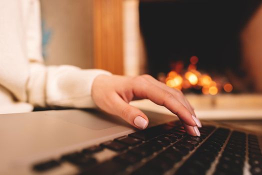 Business woman working on laptop, sitting near cozy fireplace at home. Focus on hands typing on keyboard. Social distancing, creative workspace concept. Lady shopping online through website.