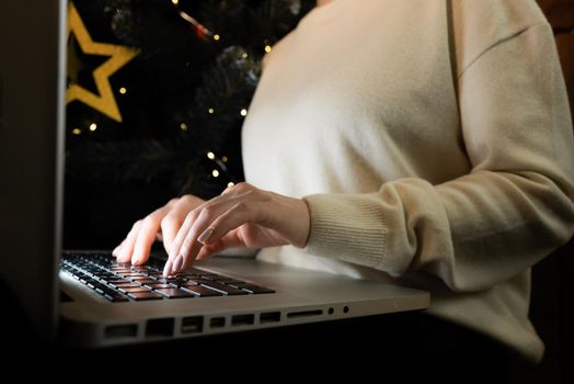 Business woman working on laptop, sitting near Christmas tree at home. Focus on hands typing on keyboard. Social distancing, creative workspace concept. Lady shopping online through website.