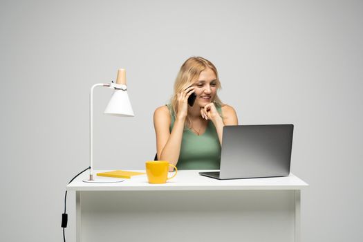 Cheerful blonde freelancer woman in green t-shirt talks on mobile phone with a clients while working on laptop in modern light office studio. Corporate Communication