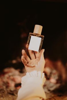 Beautiful minimalist parfume bottle in female hands on fireplace flame background. Fragrance, essence concept. High quality photo