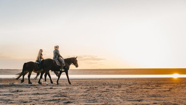 View of women riding horses along the river in golden light sunset or sunrise. Stallion walking in desert by the water