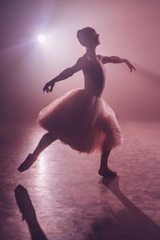 Ballet dancer in tutu performing, jumping on stage. Ballerina practices on floor in dark studio with smoke. Violet light