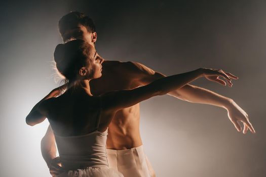 Graceful ballerina and her male partner dancing elements of classical or modern ballet in dark with floodlight backlight. Couple in smoke on black background. Art concept