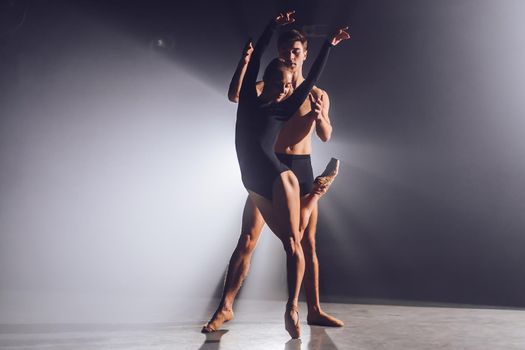 Beautiful young acrobats or gymnasts on floodlights background. Professional ballet couple dancing in spotlights smoke on big stage. Emotional duet performing choreographic art.