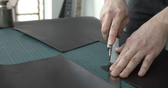 Working process of crafting a new handmade leather wallet in the leather workshop. Man's hands working with knife and iron ruler