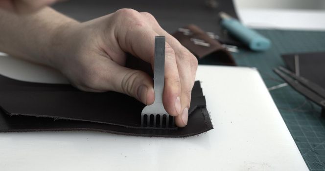 Close up men's hands make a leather product. Punching holes in the leather with a hole punch with a hammer. Tools for leather craft