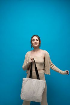 Beautiful brunette woman in a beige costume with cotton bag on in a hand on a blue background. Mockup and zero waste concept