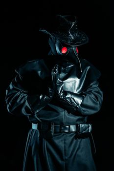 Man in plague doctor costume with crow-like mask praying with hands isolated on black background. Creepy mask, historical costume concept. Epidemic.