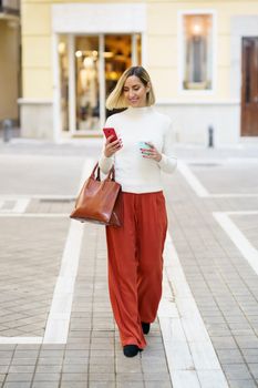 Full body of stylish female carrying handbag and cup of takeaway coffee while walking on street and using cellphone