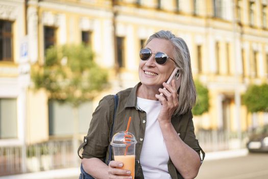 Answering phone call happy mature woman talking on the phone enjoying free time after work or traveling having juice in plastic cup on the go in city background. Enjoying life mature woman.