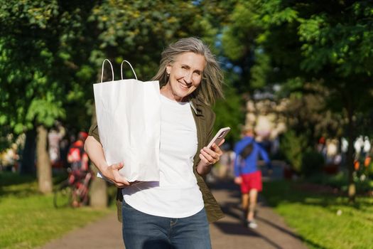 Charming mature woman walking in streets holding grocery paper bag with food and smartphone, phone in other hand cheerful smiling with blurred people life on background.