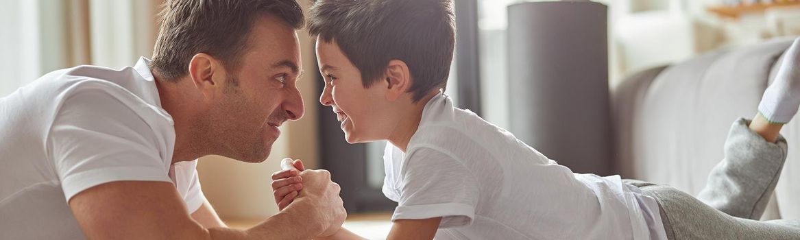 Merry man is fighting hands with little boy while having fun on floor in living room