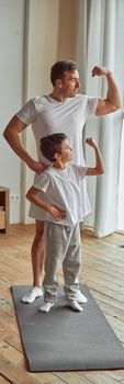 Happy muscular man and boy are standing on mat and showing biceps during domestic workout