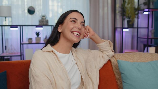 Close-up of happy calm girl in shirt smiling friendly, glad expression looking away dreaming resting, relaxation feel satisfied concept good news. Young caucasian woman sitting on sofa at home room