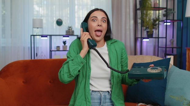 Crazy adult girl in casual shirt talking on wired vintage old-fashioned retro telephone of 80s, fooling, making silly faces. Young woman sitting on orange couch at home living room making conversation