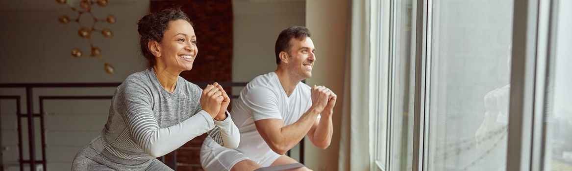 Cheerful sporty man and woman are doing squats with resistance bands before large window indoors