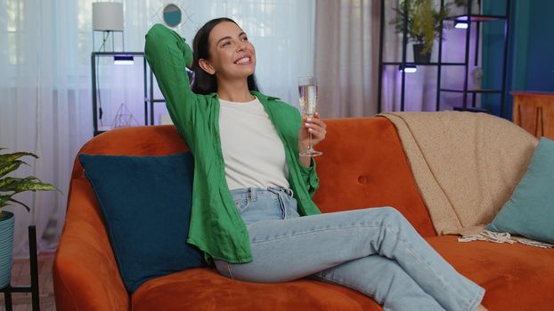 Joyful caucasian girl hold glass of champagne cheering drinking, celebrate success win, career grow up, lucky situation at home in living room. Young woman celebrating rent or purchase of new house