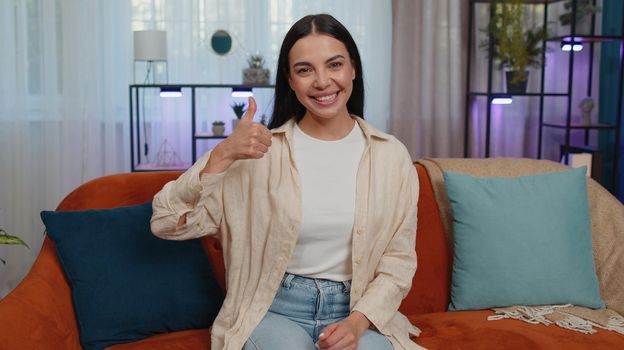 Like. Happy lovely adult girl looking approvingly at camera showing thumbs up, like sign positive something, good news, positive feedback. Young woman sitting on orange couch at home living room