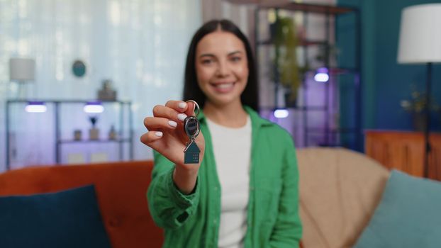 Young woman real estate agent lifting hand showing the keys of new home house apartment, buying or renting property, mortgage loan at modern home apartment indoors. Lovely girl in room sitting on sofa