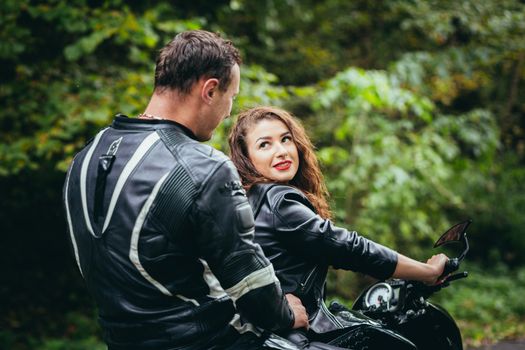 Handsome young man and beautiful young girl, couple sitting on a motorcycle in black leather clothes, close up, hugging, in nature, outdoors, looking at camera