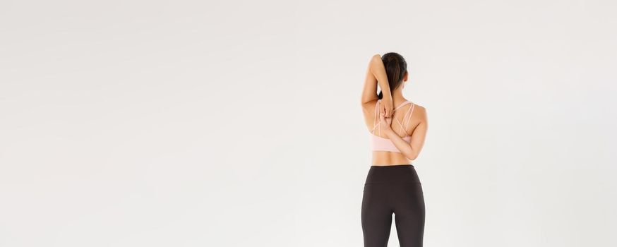 Full length rear view of active and slim brunette asian fitness girl, female athlete warm-up before yoga classes, lock hands behind back, sportswoman doing stretching exercises, white background.