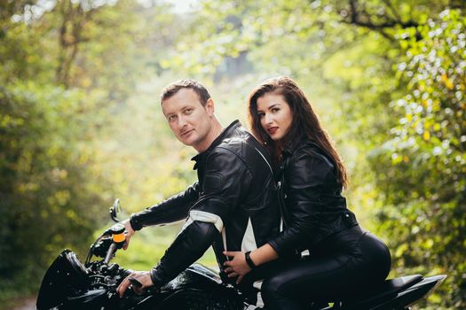 Handsome young man and beautiful young girl, couple sitting on a motorcycle in black leather clothes, close up, hugging, in nature, outdoors, looking at camera