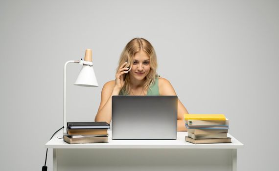 Young attractive business woman with a blond hair sitting at the table and working on a laptop and talking with a client on the phone. Young cheerful student girl talking with a smartphone. Studying