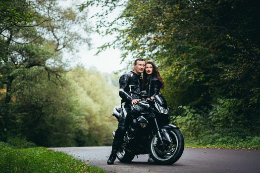 Handsome young man and beautiful young girl, couple sitting on a motorcycle, standing near a motorcycle in black leather clothes, hugging, in nature, on the street