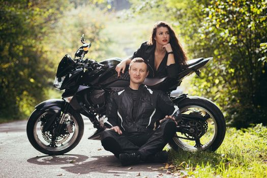 Handsome young man and beautiful young girl, couple sitting near motorcycle in black leather clothes, hugging, in nature, outdoors