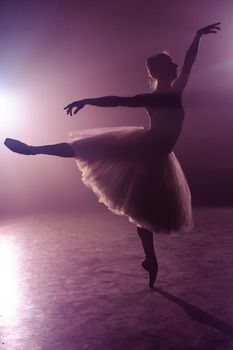 Ballet dancer in tutu performing, jumping on stage. Ballerina practices on floor in dark studio with smoke. Violet light