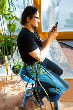 a man works at a phone in a home office, sitting on a Orthopedic ergonomic kneeling chair on a glazed balcony among greenery. back health care
