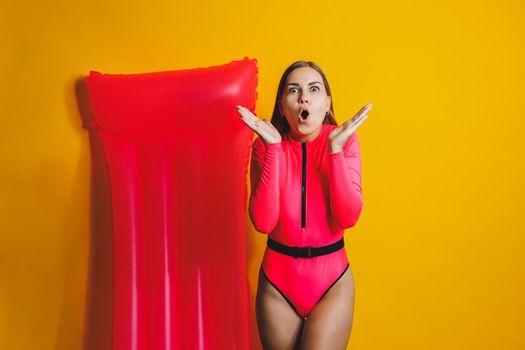 Beautiful young smiling woman wearing a pink swimsuit and posing on a yellow background with an air mattress, isolated on a bright yellow background