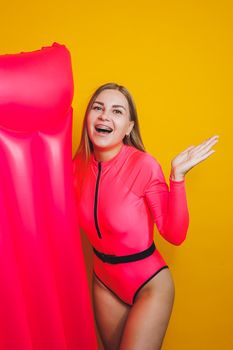 Young slender woman in a pink swimsuit on a yellow background. Girl with an inflatable mattress for swimming in the pool. Summer vacation.
