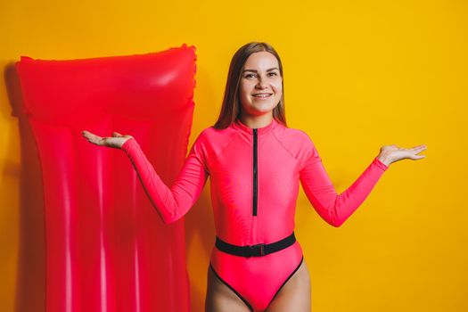 Beautiful young smiling woman wearing a pink swimsuit and posing on a yellow background with an air mattress, isolated on a bright yellow background