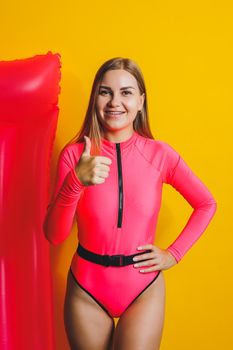 Beautiful young smiling woman wearing a pink swimsuit and posing on a yellow background with an air mattress, isolated on a bright yellow background