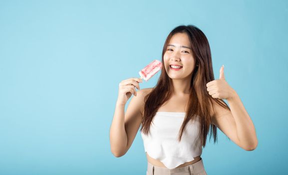 Sweet tasty frozen dessert icecream on summer time. Happy Asian young beautiful woman holding delicious ice cream wood stick mixed fruit flavor and show thumb up good sign, isolated on blue background