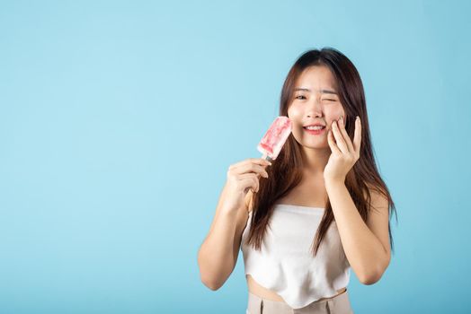 Portrait of Asian young woman with sensitive teeth after eating delicious ice cream wood stick mixed fruit flavor feeling painful uncomfortable, studio shot isolated on blue background, dental problem