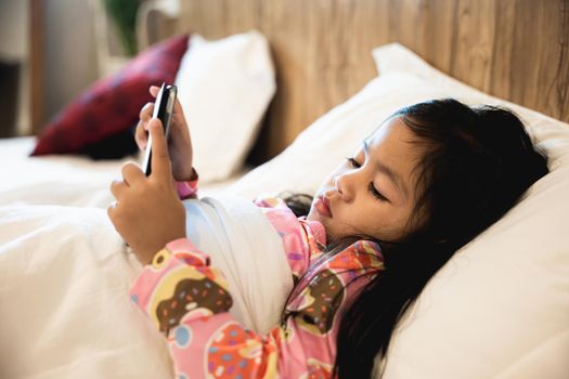 Little Asian girl playing digital tablet on bed addicted game and cartoon, Kid watching videos and cartoons online in bedroom at home, Fun online concept