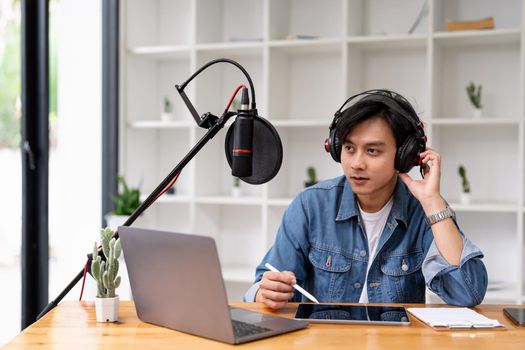 Asian man recording a podcast on laptop computer with microphone while online live streaming at home.