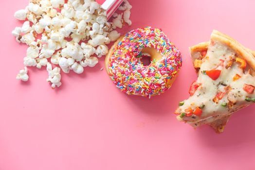 Slice of cheese pizza on a plate , donuts and popcorn on pink ,